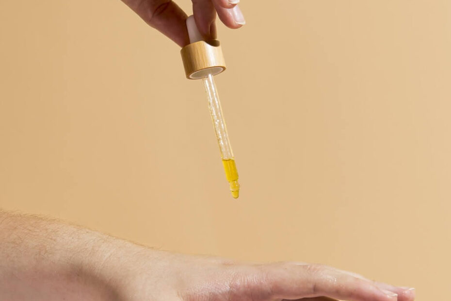 Ręka trzyma zakraplacz nad drugą ręką, dozując żółty płyn na skórę, na beżowym tle.