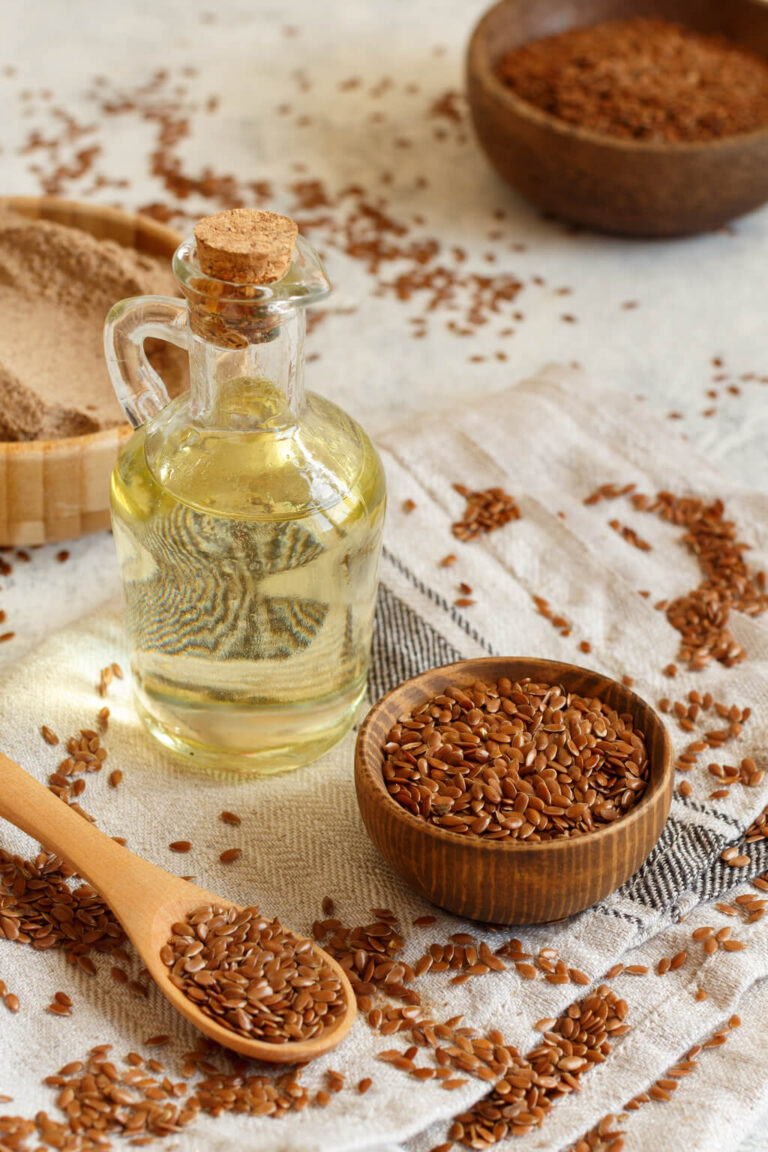 Szklana butelka naturalnego oleju lnianego zimnotłoczonego z korkiem stoi na ściereczce obok drewnianej miski i łyżki wypełnionej brązowymi nasionami lnu. Luźne nasiona lnu są rozsypane na ściereczce i otaczającej ją powierzchni.