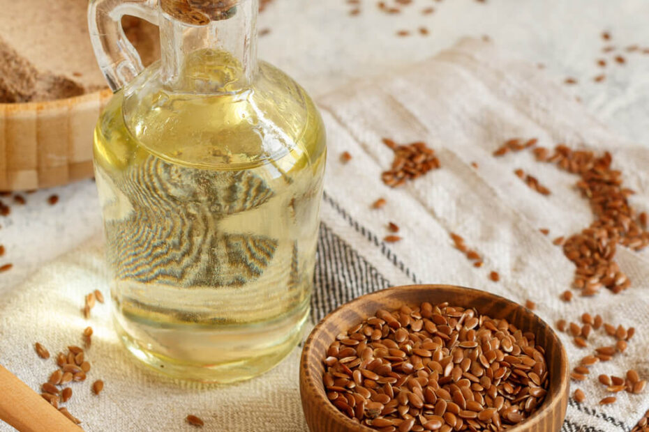 Szklana butelka naturalnego oleju lnianego zimnotłoczonego z korkiem stoi na ściereczce obok drewnianej miski i łyżki wypełnionej brązowymi nasionami lnu. Luźne nasiona lnu są rozsypane na ściereczce i otaczającej ją powierzchni.