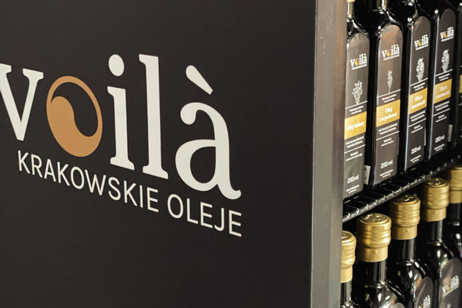 Zbliżenie na czarną witrynę z napisem voilà KRAKOWSKIE OLEJE obok półek z butelkami oleju z czarnymi etykietami i złotymi nakrętkami.