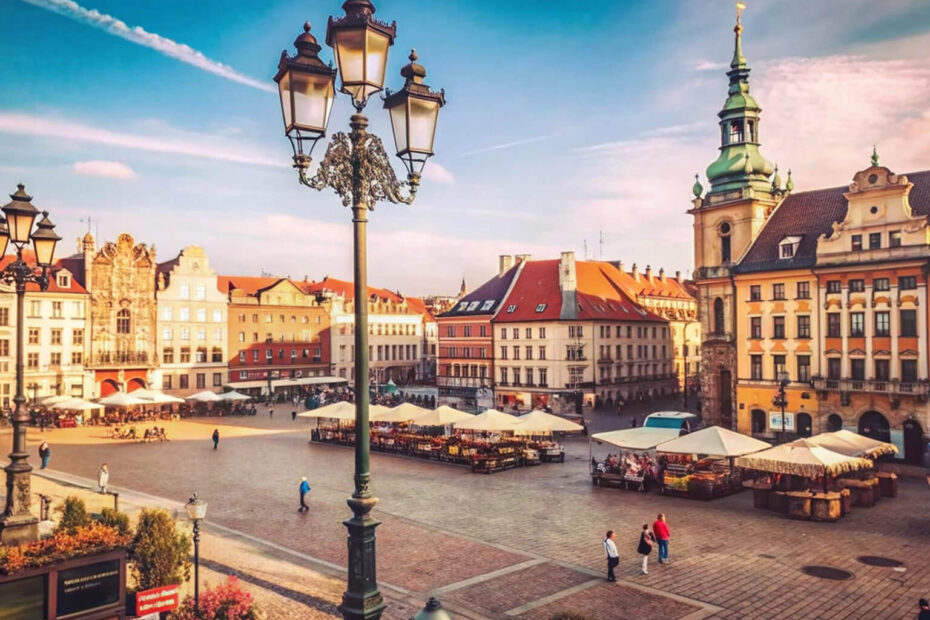 Duży europejski rynek z zabytkowymi budynkami, wysoką wieżą zegarową, latarniami ulicznymi i straganami z białymi parasolami. Ludzie spacerują i cieszą się słonecznym dniem pod błękitnym niebem z lekkimi chmurami.