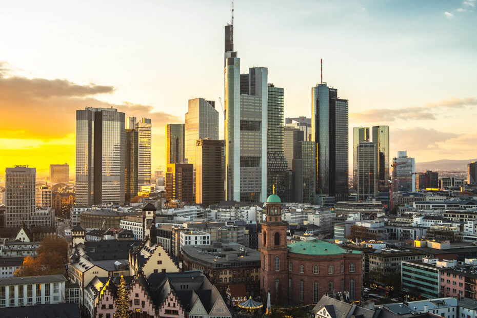 Panorama Frankfurtu o zachodzie słońca, z nowoczesnymi drapaczami chmur, zabytkowymi budynkami na pierwszym planie i ciepłym pomarańczowym niebem z rozproszonymi chmurami.